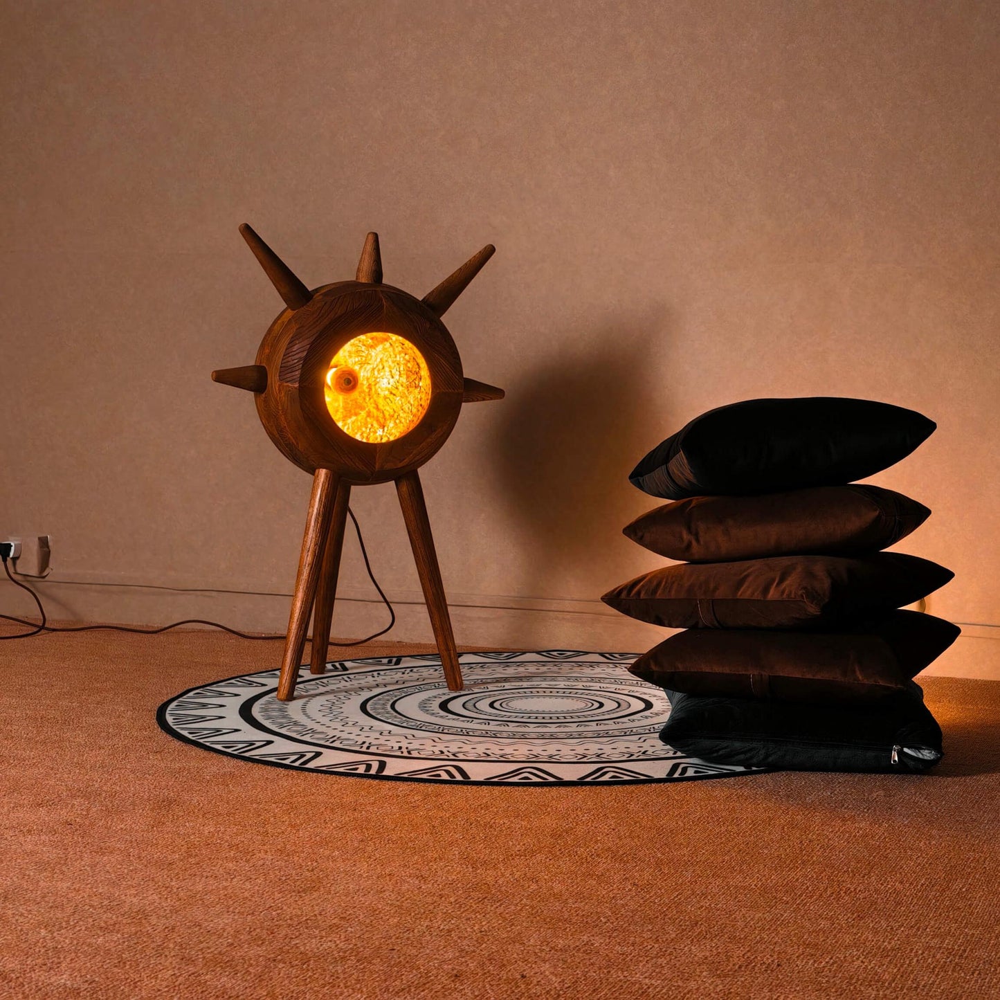 Wooden lamp with a glowing light bulb on a patterned rug, surrounded by stacked pillows against a plain wall.